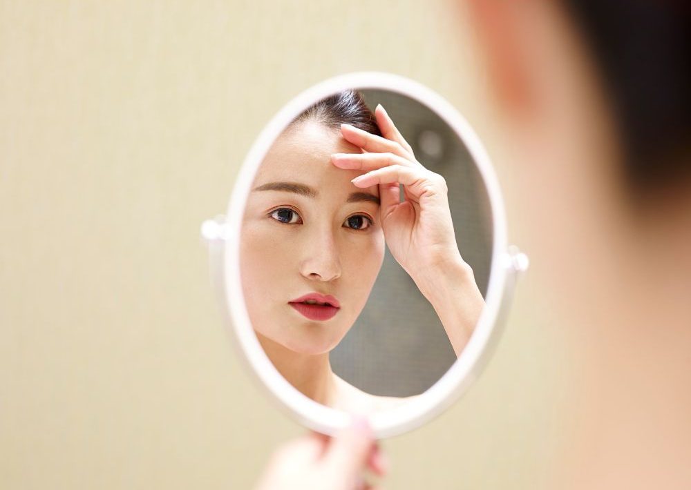 Young Asian woman looking at self in mirror - imtmphoto - Canva - Getty Images